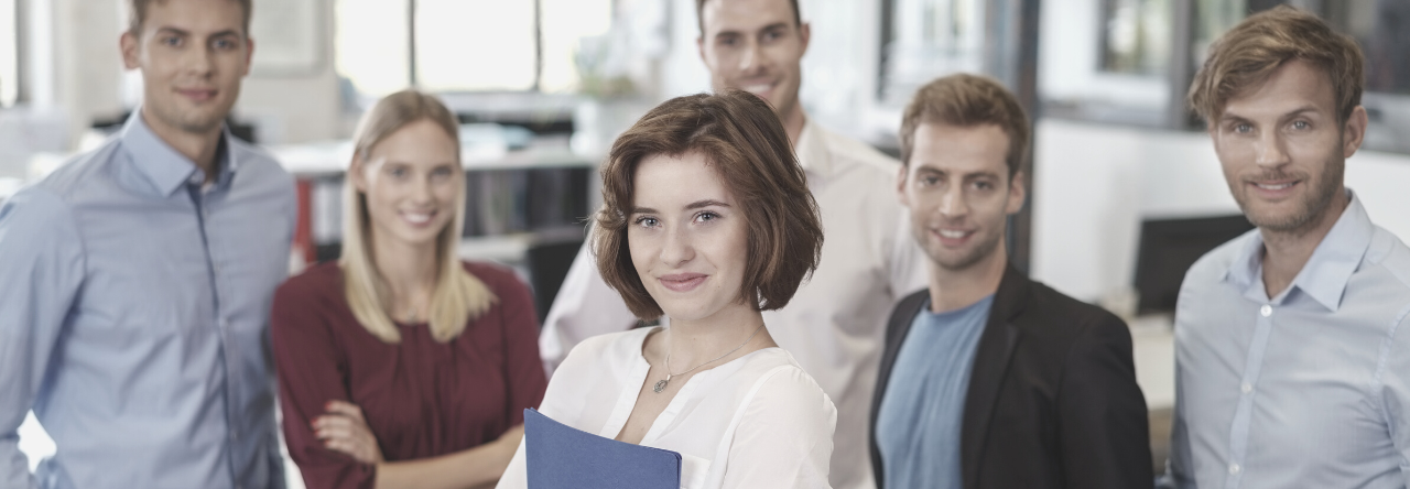 Eine Gruppe von sechs Personen steht zusammen in einem modernen Büro. Eine Frau in einem hellen Oberteil hält einen blauen Ordner in der Hand und lächelt. Die anderen sind in formeller Kleidung und wirken freundlich. Sie repräsentieren Teamarbeit und Professionalität.